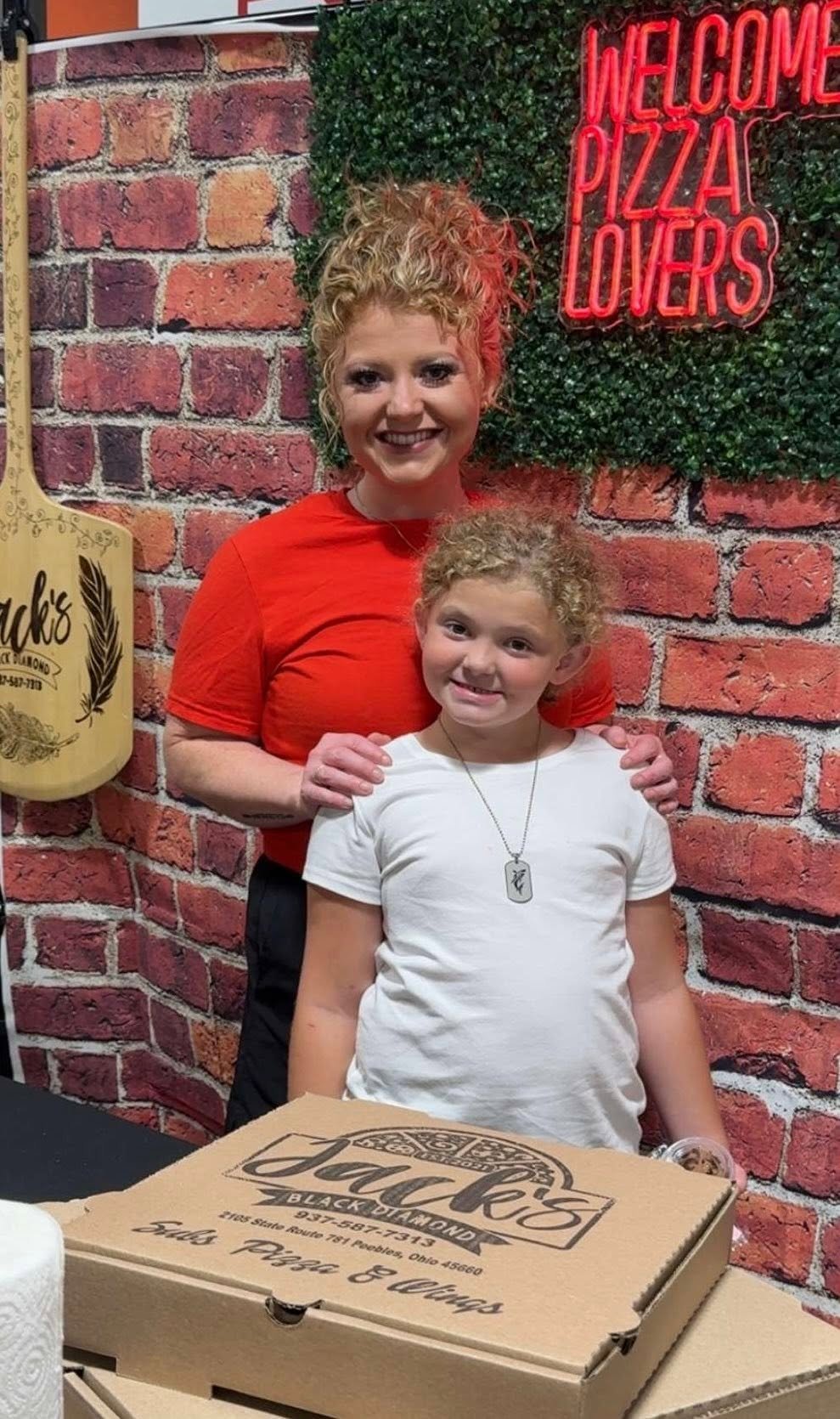 Jacqueline, owner of Local Jack's, smiling with her daughter in front of the restaurant's welcome sign